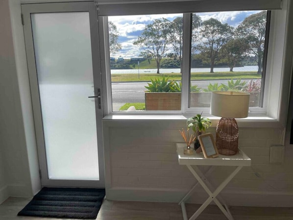 A room with a view of a lake and trees, a white door, a small table with plants and a candle holder, and a window with a view of a road and greenery.