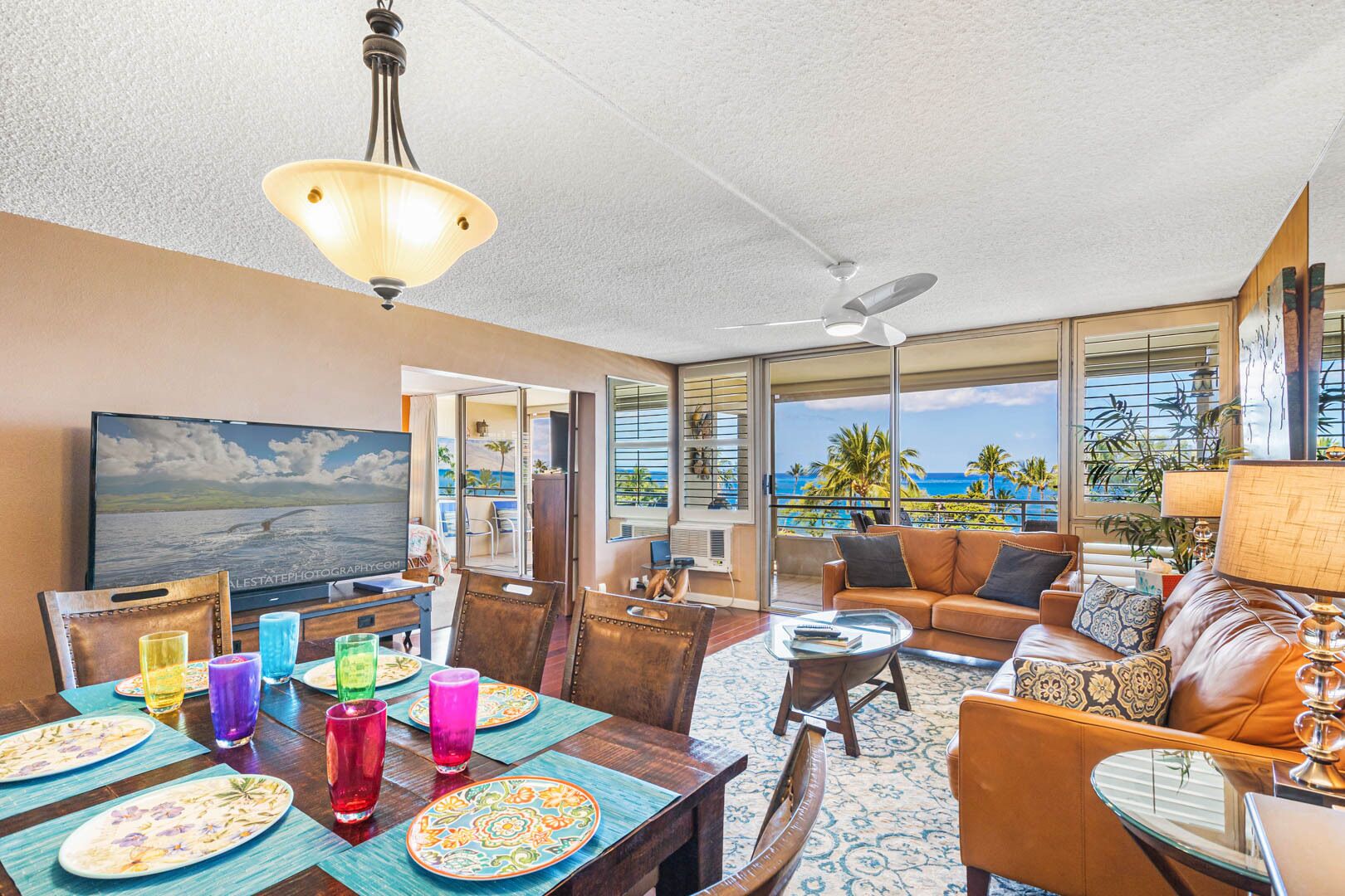 A spacious living area with a large TV, dining table set with colorful plates and glasses, a sofa, and a balcony with a view of the ocean.