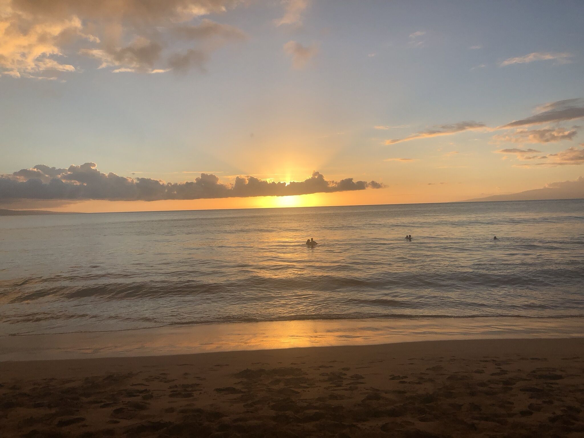 A beach scene with the sun setting over the ocean, calm waters, and footprints on the sand.