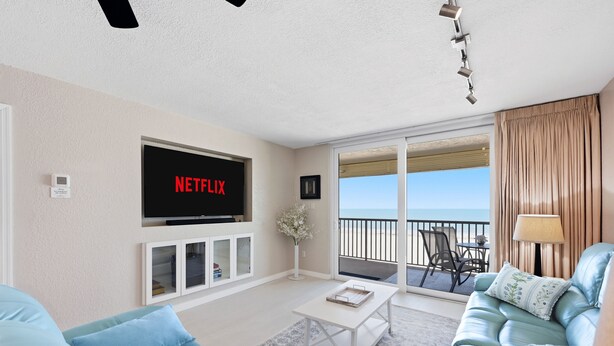 A living room with a TV displaying 'NETFLIX', a sofa, a coffee table, and a view of the ocean through a balcony.
