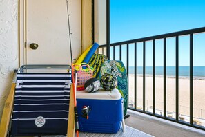 Un balcón con vistas a la playa, un enfriador azul, un bolso a rayas, una cesta colorida y una almohada estampada.
