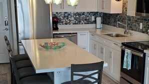 Une cuisine moderne avec des meubles blancs, un réfrigérateur en acier inoxydable, une table à manger avec des chaises et des luminaires suspendus.