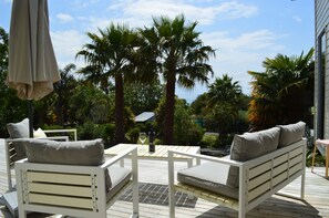 A deck with white furniture, including a sofa and chairs, surrounded by palm trees and a view of the ocean. - Luxurious Holiday Home (Mangonui)