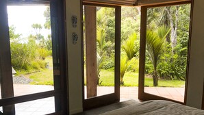 A bedroom with a large bed, a view of a lush garden through open sliding doors, and a ceiling fan.