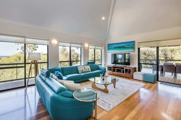 A spacious living room with a teal sofa, a glass coffee table, and a television on a wooden stand.