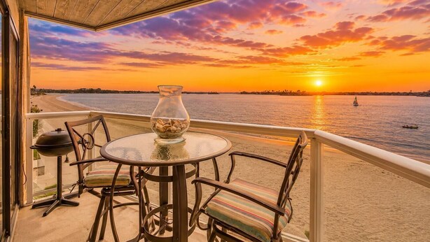 Uma varanda com vista para o pôr do sol sobre a água, uma mesa com cadeiras e um churrasqueira.