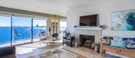 A spacious living room with a fireplace, a television, and a balcony with a view of the water.