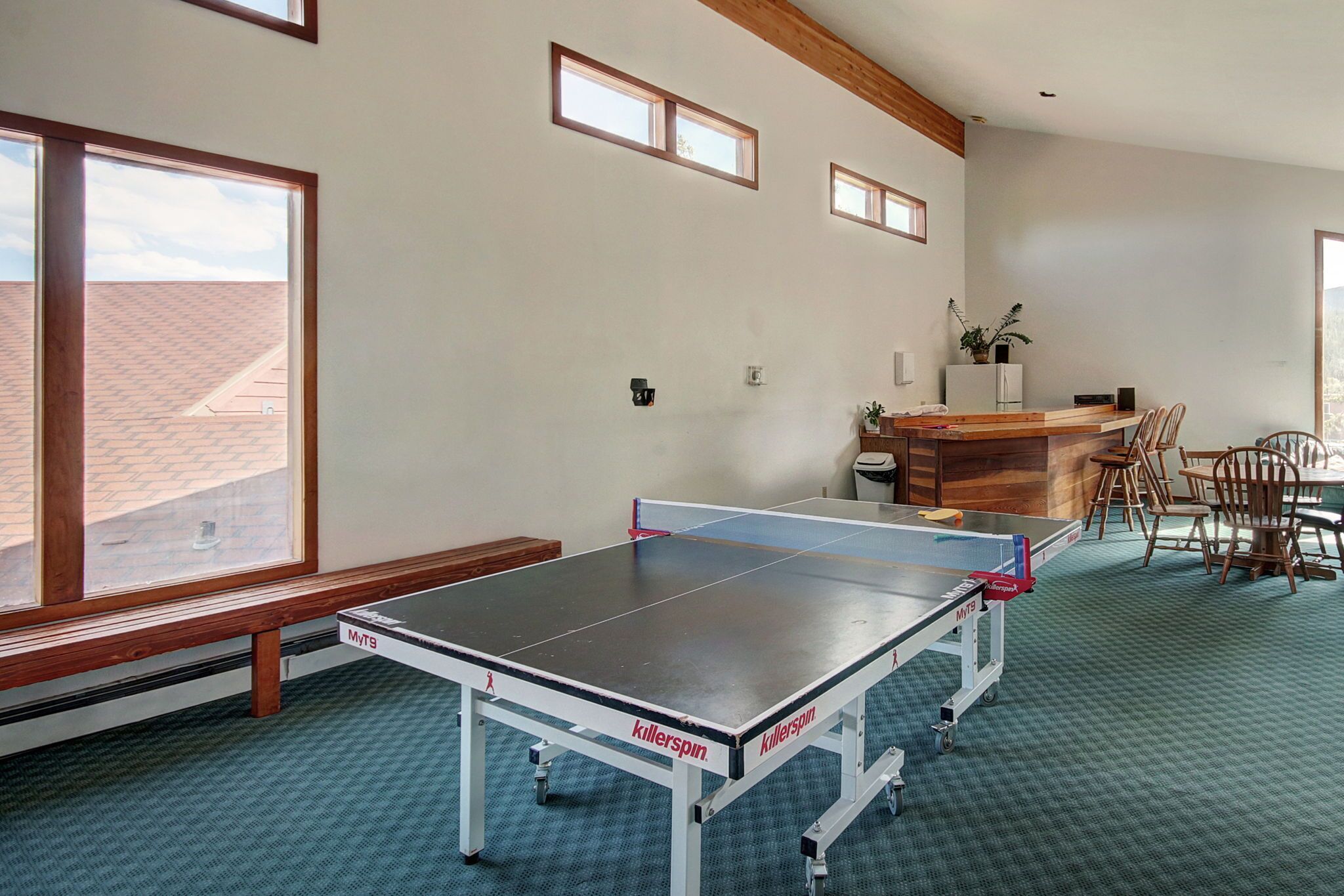 Una mesa de ping pong en una habitación con ventanas amplias, techo de madera y una zona de cocina con sillas.