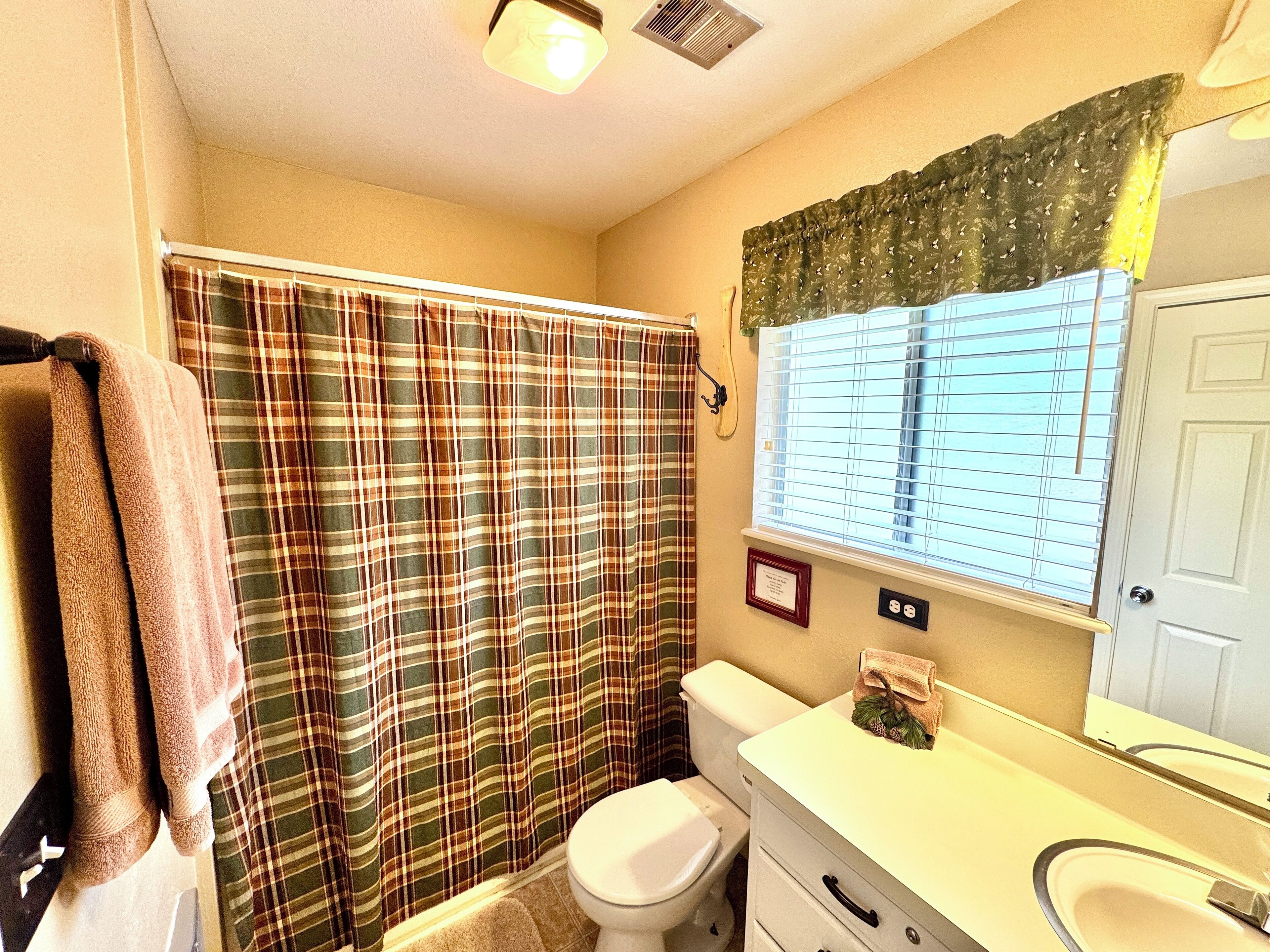 A bathroom with a plaid shower curtain, a white toilet, a vanity with a mirror, and a window with blinds.