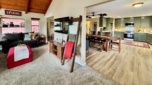 Amplia sala de estar con techo de madera, un pouf rojo, un sofá y un televisor. Se ve un comedor con mesa y sillas de madera. En el fondo, una cocina con armarios verdes y un microondas.