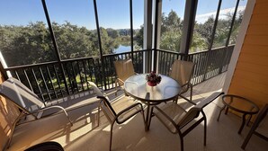 Un balcon avec une table ronde et des chaises, un vase de fleurs et une vue sur des arbres et un lac.