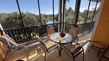 Un balcon avec une table ronde et des chaises, un vase de fleurs et une vue sur des arbres et un lac.
