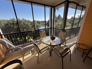 Un balcone con un tavolo rotondo e sedie, un vaso di fiori e vista su alberi e un lago.