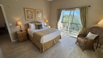 Une chambre avec un grand lit, deux lampes de chevet, un fauteuil en osier et un balcon avec des rideaux.