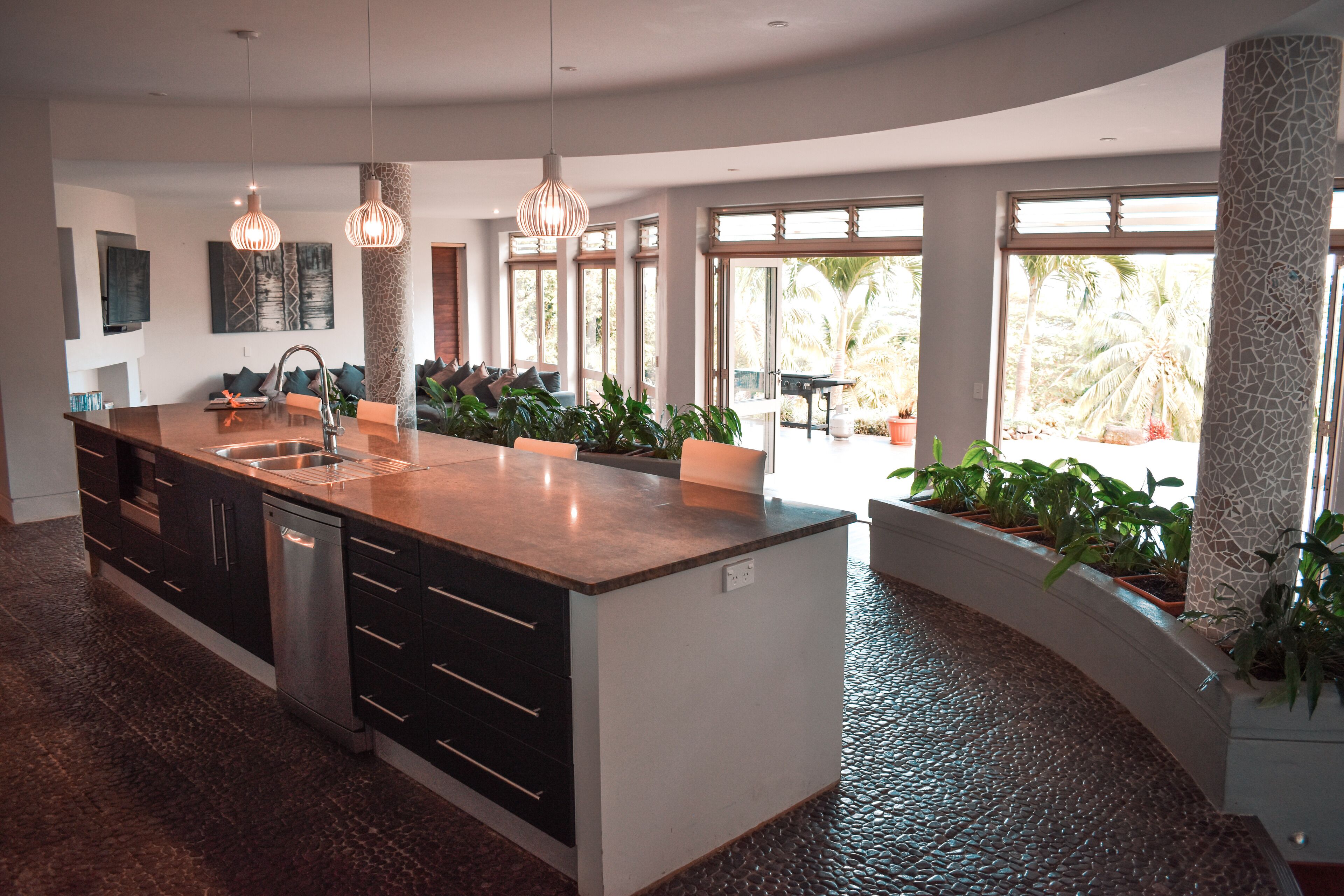 A modern kitchen with a large island, stainless steel appliances, pendant lighting, and large windows offering views of the outdoors.
