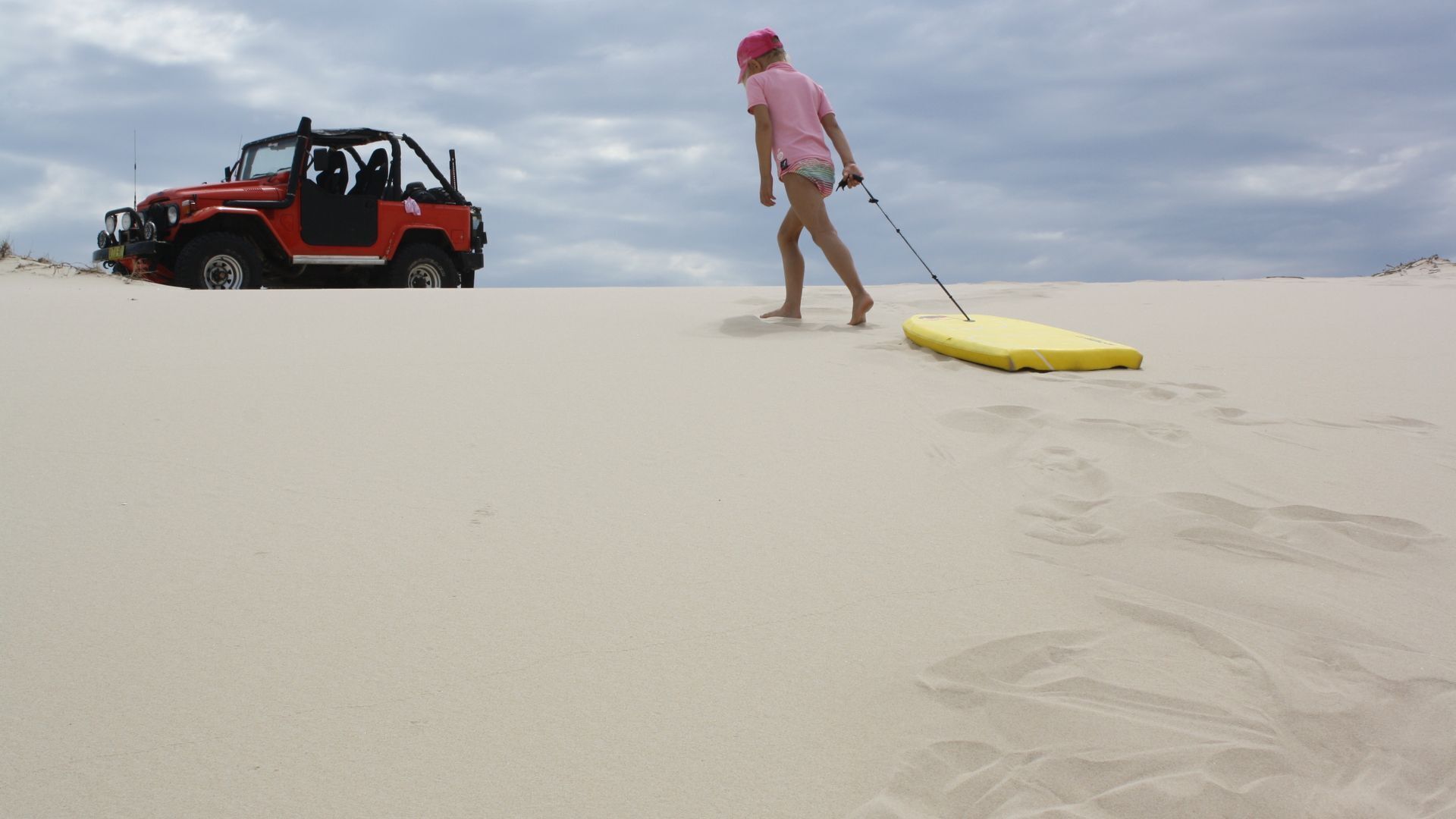 A person walking on a sandy beach with a yellow surfboard and a red off-road vehicle in the background.