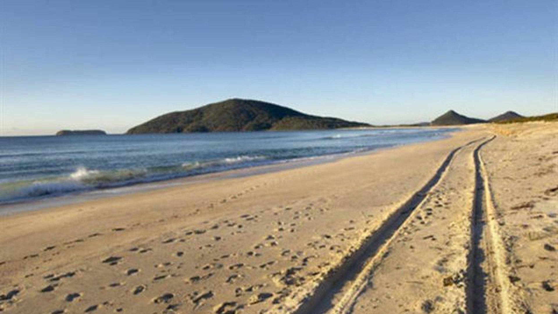 A sandy beach with a clear path leading to the water, a distant mountain, and calm waves.