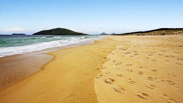 A sandy beach with footprints and a clear view of the ocean and distant hills.