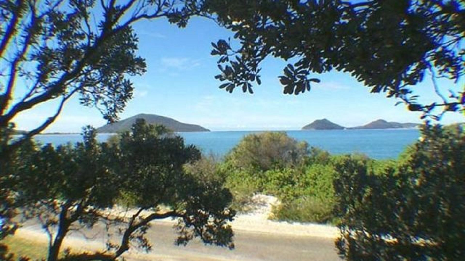 Uma praia com árvores em primeiro plano e uma vista clara do mar e ilhas distantes.