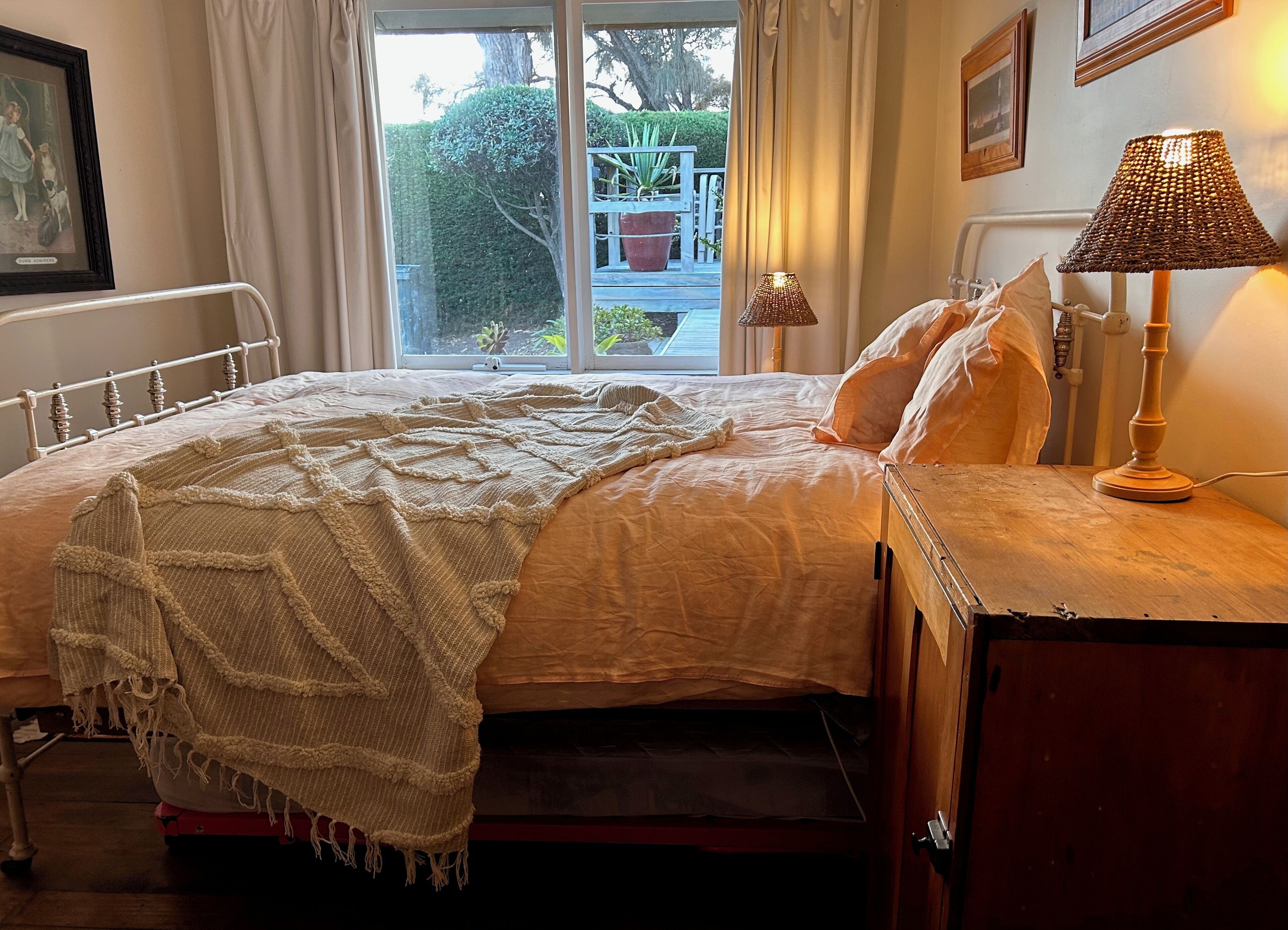A neatly made bed with a white blanket and pillows, a bedside table with a lamp, and a window with curtains.