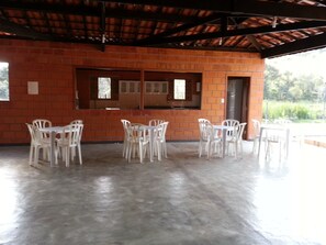 A covered outdoor dining area with brick walls and a concrete floor, featuring white plastic chairs and tables. - Chacara Dois Lagos!   Come and admire nature in this place!   (ITU)