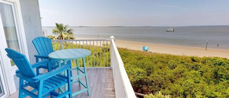 A balcony with blue Adirondack chairs and a small table, overlooking a beach with a boat in the distance.