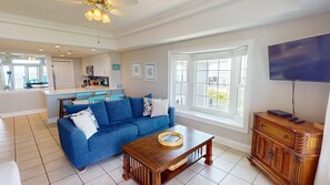 Un salon spacieux avec un canapé bleu, une table basse en bois et un téléviseur posé sur un meuble en bois. On aperçoit une cuisine à l’arrière-plan, avec vue sur un balcon.