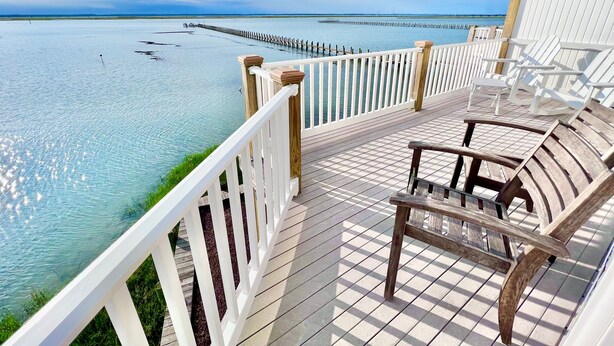 Una terraza de madera con barandas blancas da vista a un mar tranquilo, con una fila de sillas y un muelle distante.