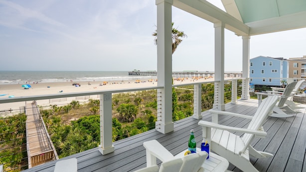 Une terrasse couverte, avec des chaises et des tables blanches, donnant sur une plage où l’on aperçoit une jetée et des bâtiments en arrière-plan.