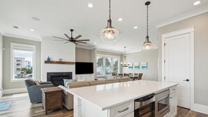 Une cuisine moderne dotée d’un grand îlot central, de meubles blancs et d’un four encastré. On y trouve également un ventilateur de plafond, un éclairage encastré et une vue sur l’extérieur grâce à de grandes fenêtres.