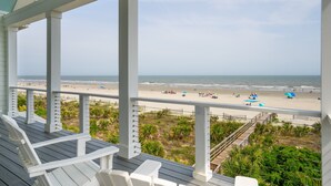 Une terrasse couverte, avec des chaises blanches, donnant sur une plage parsemée de parasols et fréquentée par des gens.