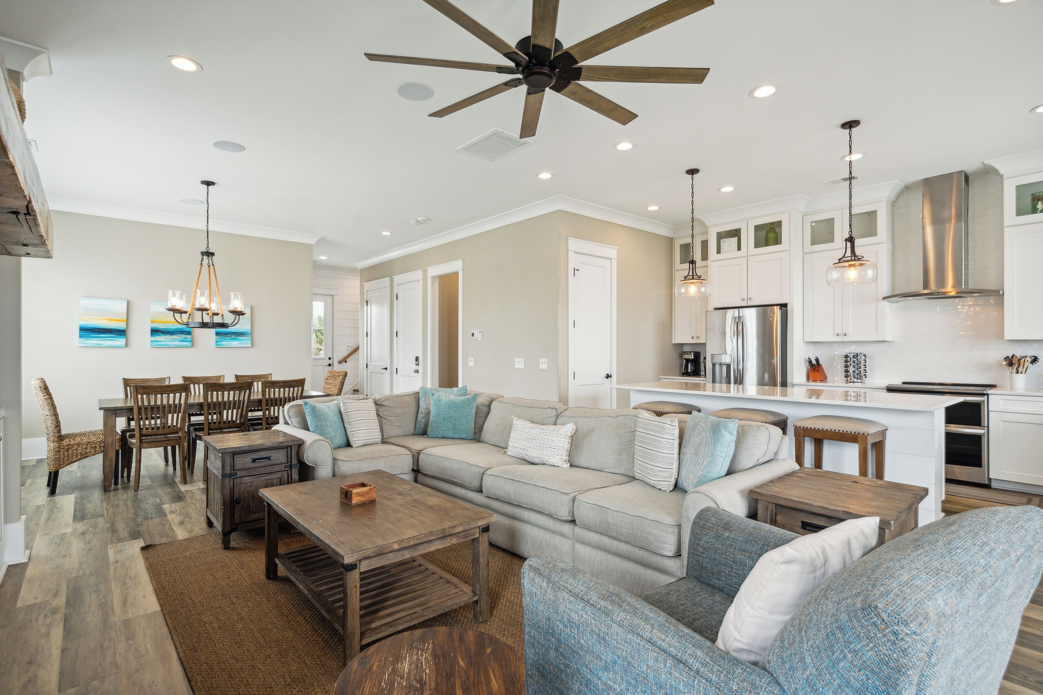 A spacious living area with a large sofa, a wooden coffee table, and a dining table with chairs. There is a kitchen area with white cabinets and stainless steel appliances.