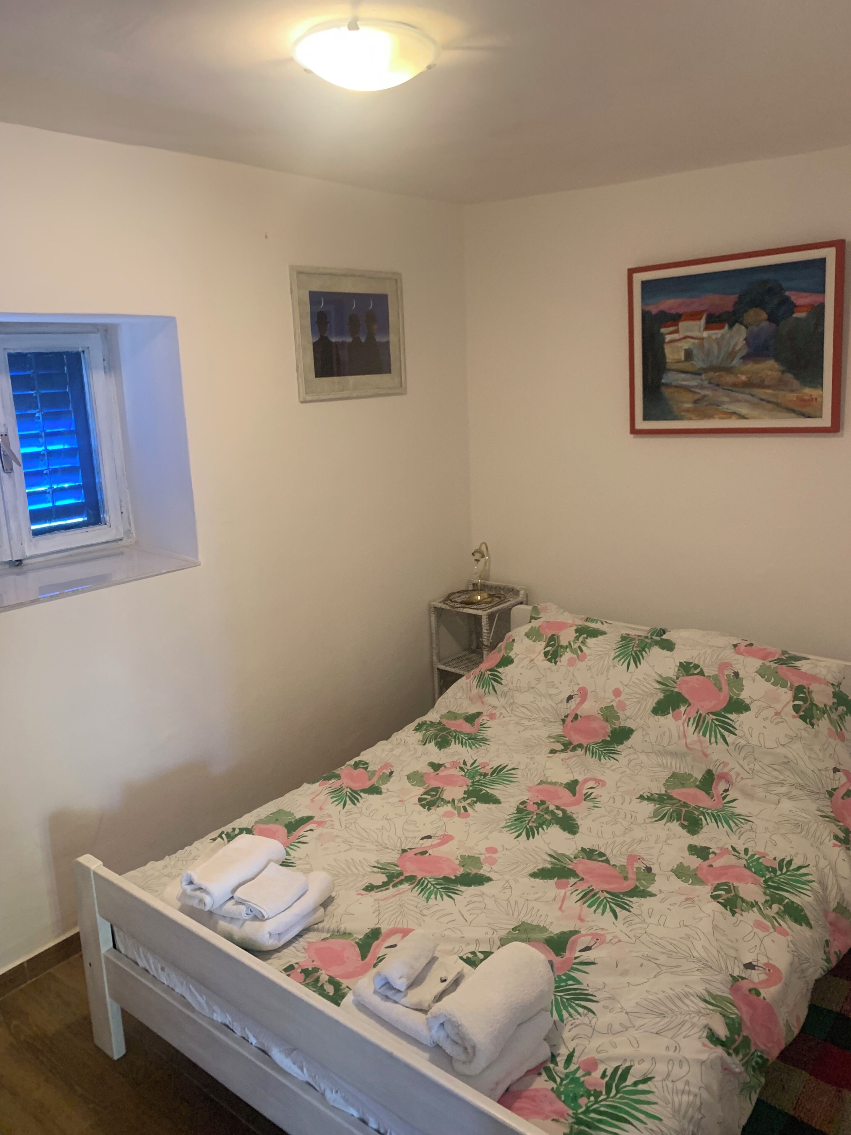 Ein Schlafzimmer mit einem Bett, einem Fenster mit Jalousien, einem gerahmten Bild und einem Nachttisch mit Lampe.