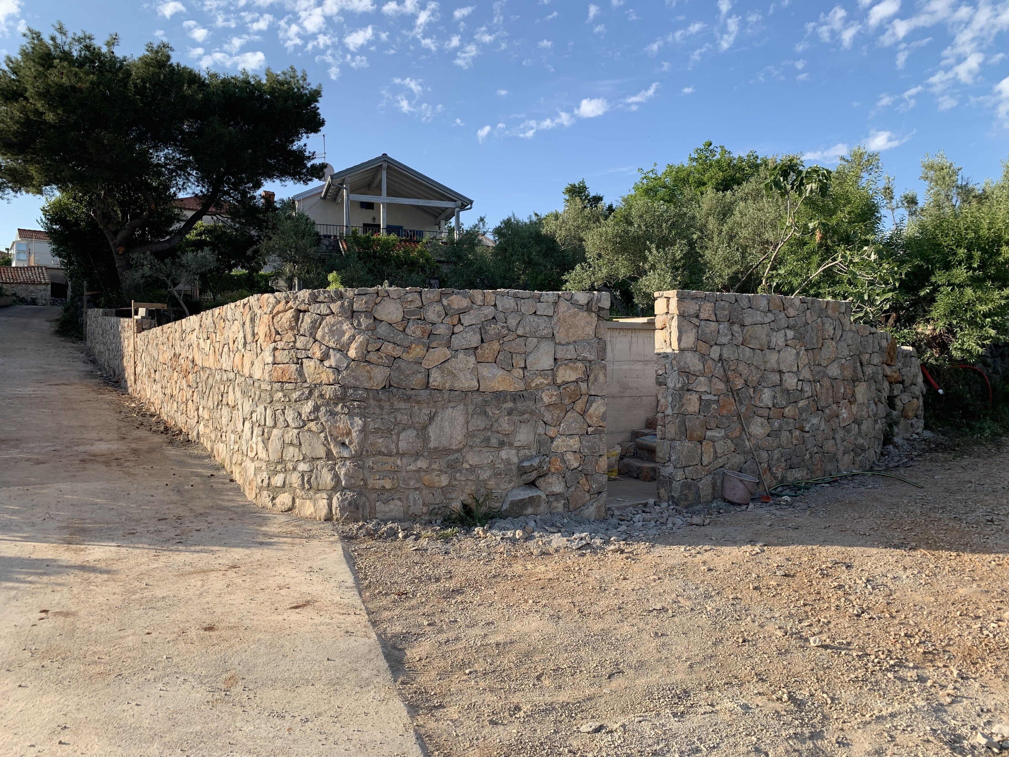 Estructura de pared de piedra con techo plano, rodeada de árboles y una casa al fondo.