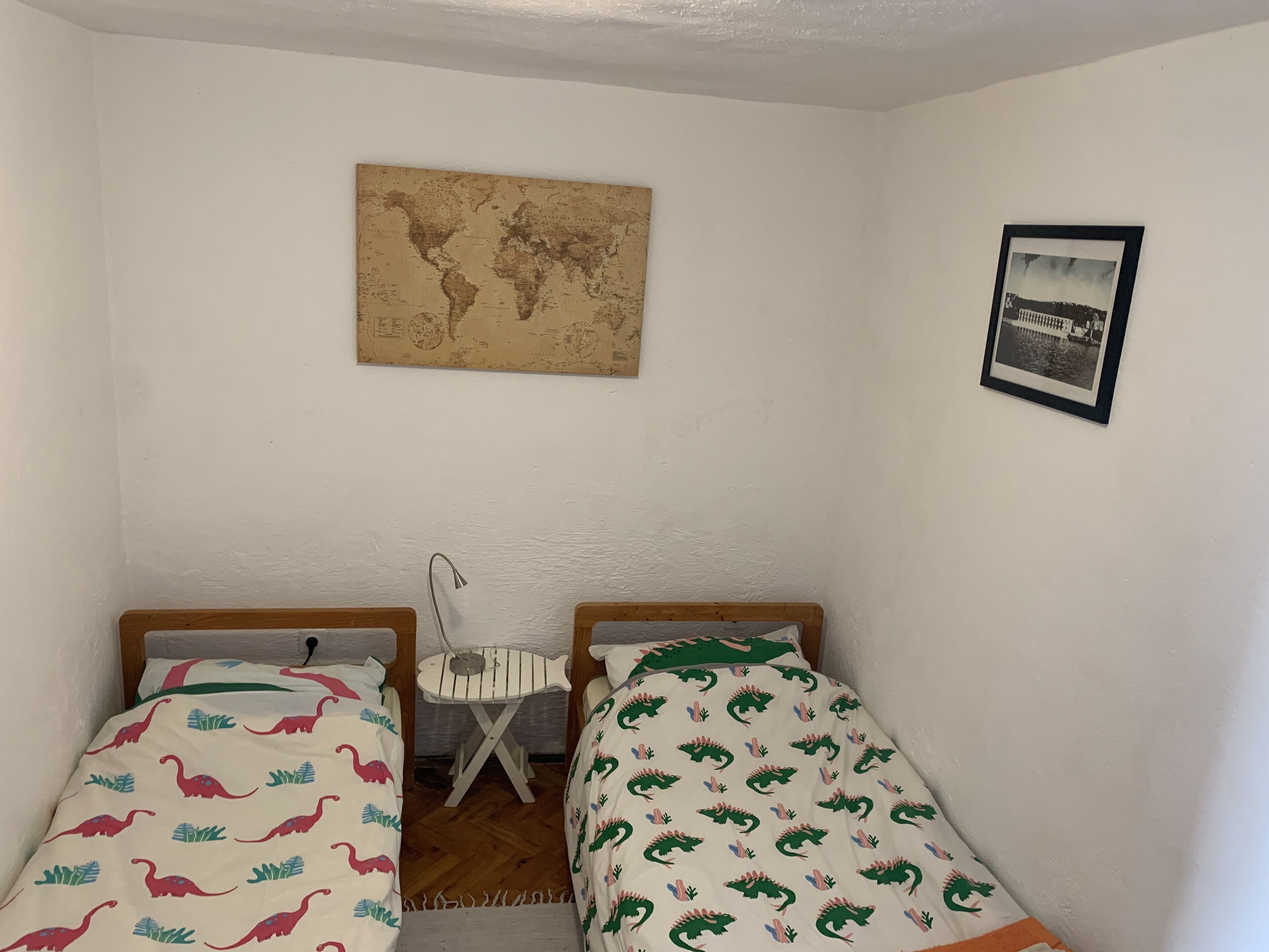 Una habitación pequeña con dos camas individuales, una mesita, una silla, un mapa del mundo en la pared y un cuadro enmarcado.