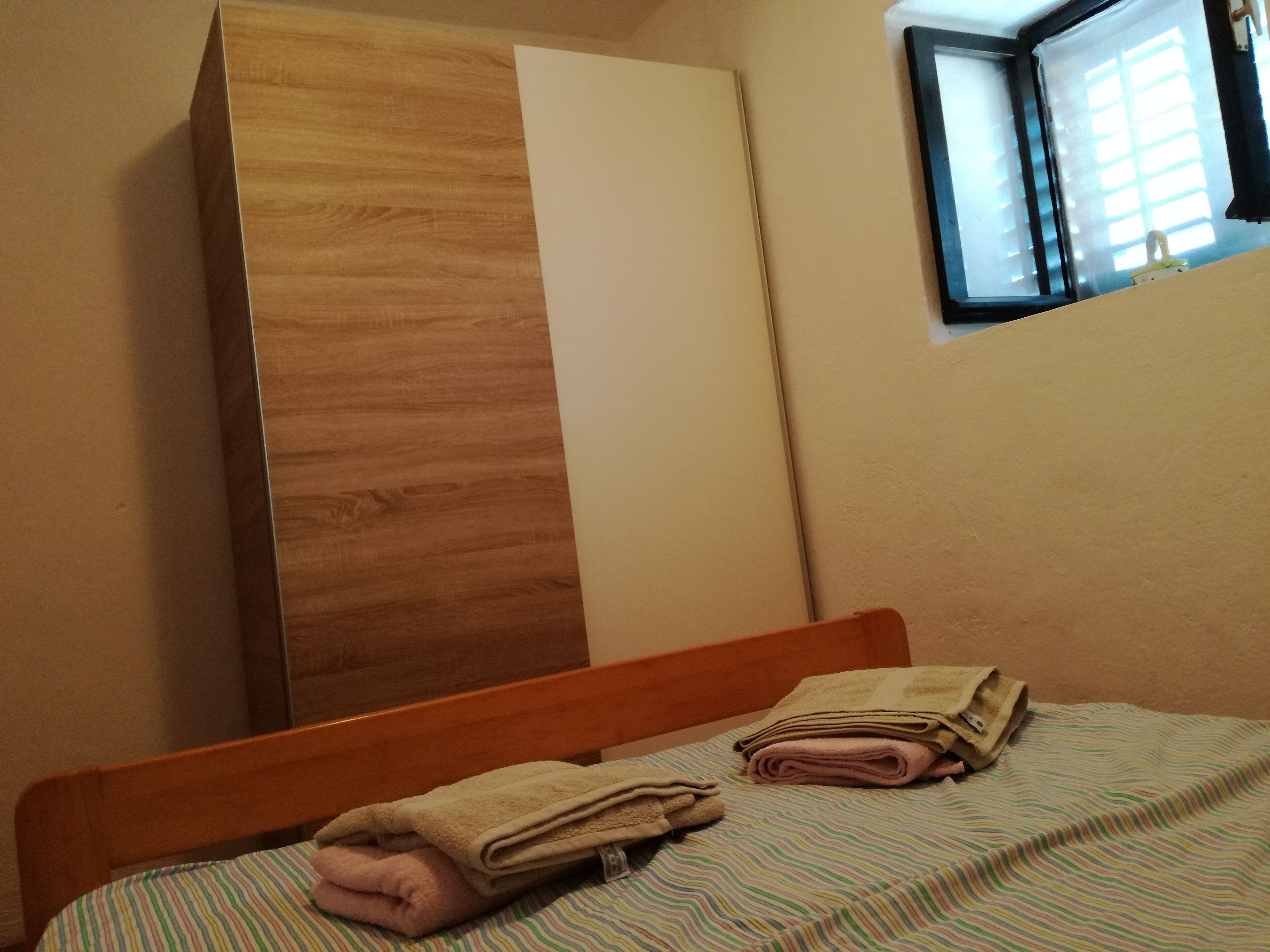 A bedroom with a wooden wardrobe, a bed with striped bedding, and a window with blinds.
