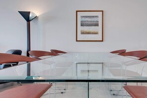 Un salón de conferencias con una mesa de cristal, seis sillas marrones, un cuadro de un paisaje enmarcado y una lámpara de pie alta.