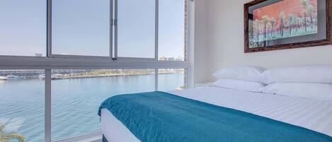 Habitación de hotel con un ventanal grande que ofrece vistas a un cuerpo de agua y un puente. La habitación tiene una cama con ropa blanca y una manta color turquesa.