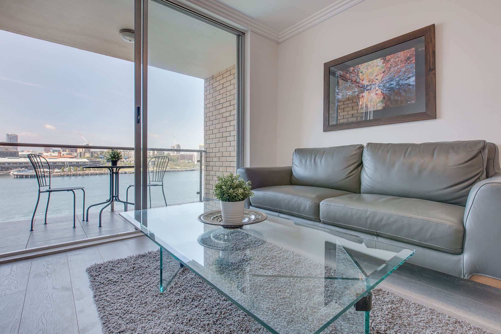 Ein modernes Wohnzimmer mit Glastisch, grauem Sofa und Blick auf die Stadt durch große Fenster.
