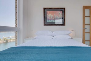 Ein Schlafzimmer mit einem Bett, weißen Kissen, einer blauen Decke, einem Nachttisch mit einer Lampe und einem Bild an der Wand.