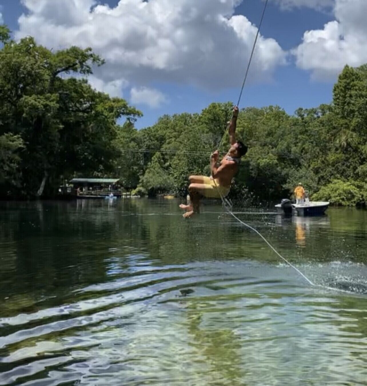A person ziplining over a body of water with trees and a boat in the background.