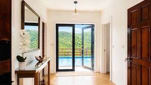 L’entrée d’un hôtel, avec un plancher en bois, une porte en verre donnant sur un balcon avec une piscine et une vue sur une colline boisée.