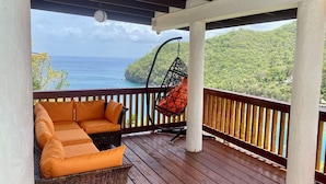Une terrasse en bois avec un canapé en osier et un fauteuil suspendu, offrant une vue sur un paysage côtier.