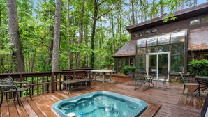 Una terraza de madera con un jacuzzi, rodeada de árboles y un área de patio con paredes de vidrio.