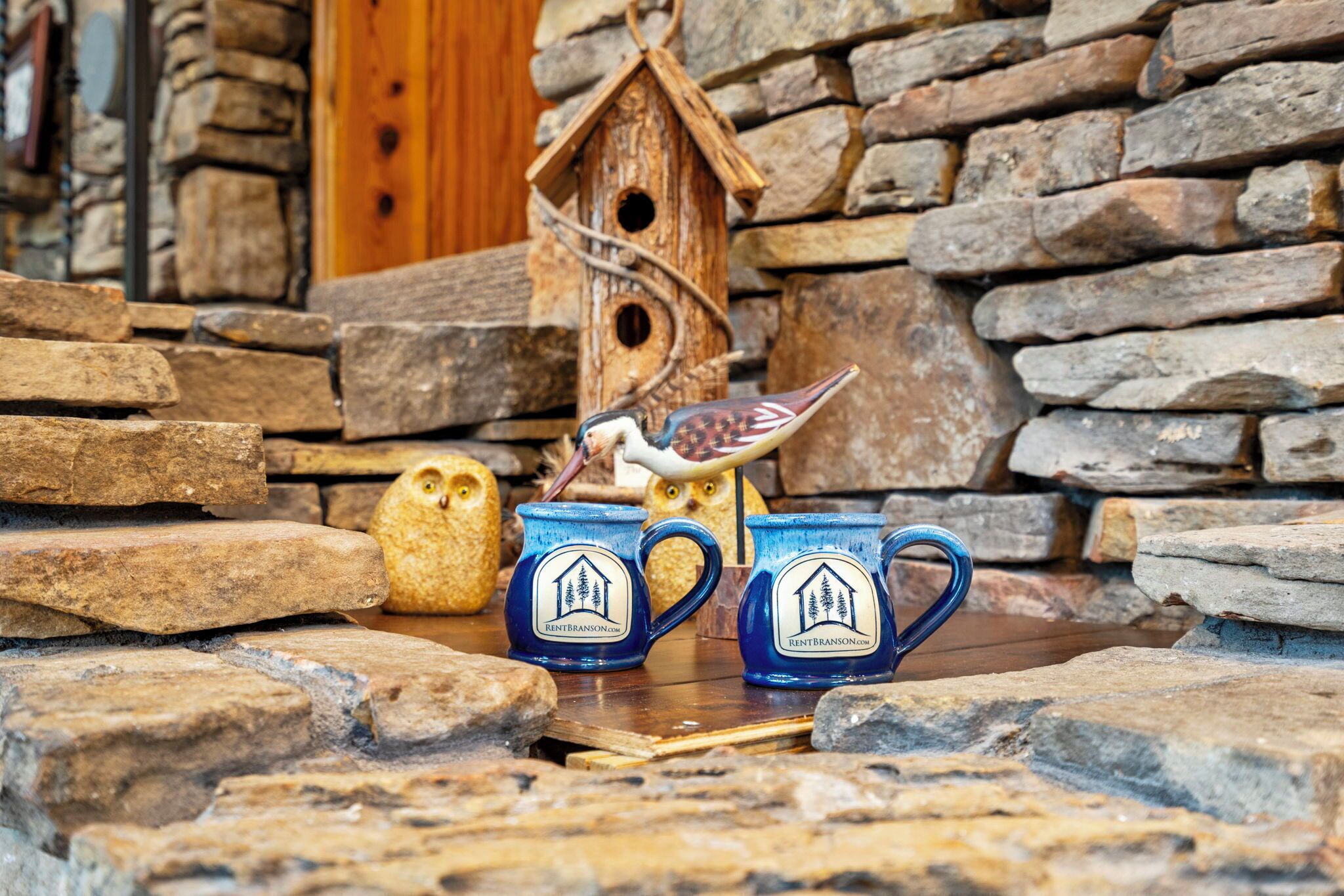 Een rustieke omgeving met stenen muren, een houten vogelhuisje en twee keramische mokken met een logo van een huis.