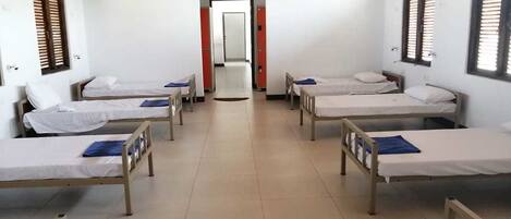 Habitación tipo dormitorio con cuatro camas individuales alineadas, suelo de baldosas y una puerta al fondo.