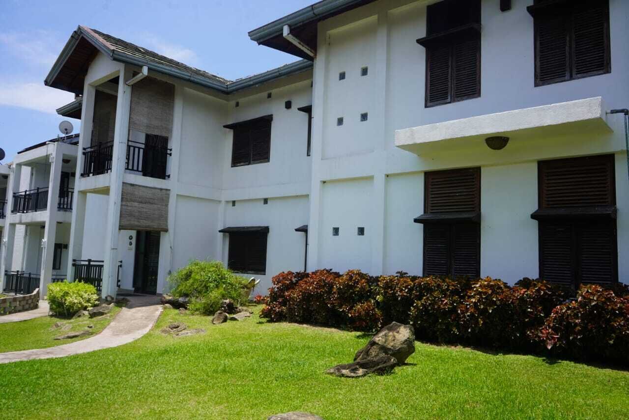 A multi-story building with a white facade, balconies, and a well-maintained lawn with shrubs and rocks.