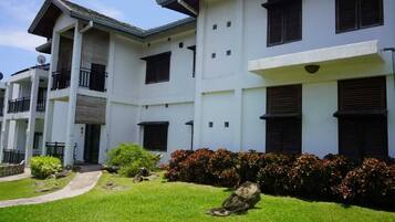 A multi-story building with a white facade, balconies, and a well-maintained lawn with shrubs and rocks.