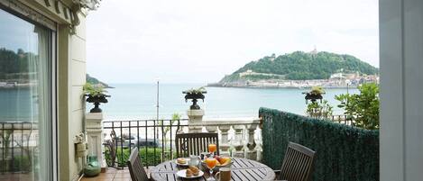 A balcony with a table set for breakfast, overlooking a coastal view with a hill and cityscape.