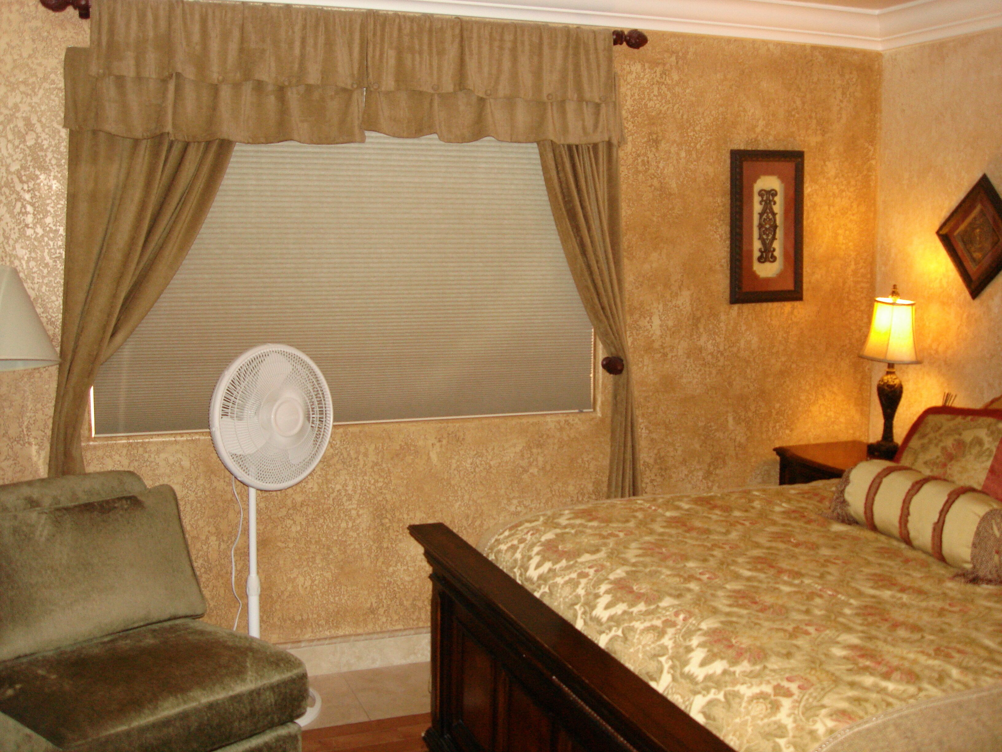 A bedroom with a bed, a chair, a standing fan, and a nightstand with a lamp.